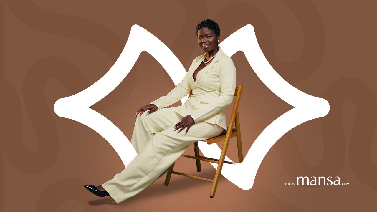Mansa Richardson in a cream suit and pearls, seated on a wooden chair in front of the This is Mansa mark